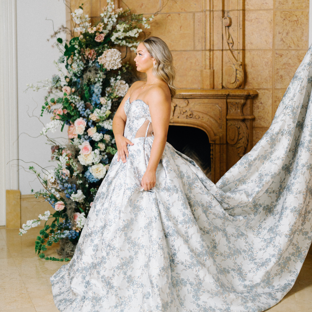 Bride in silver floral ballgown with flowing train beside ornate fireplace