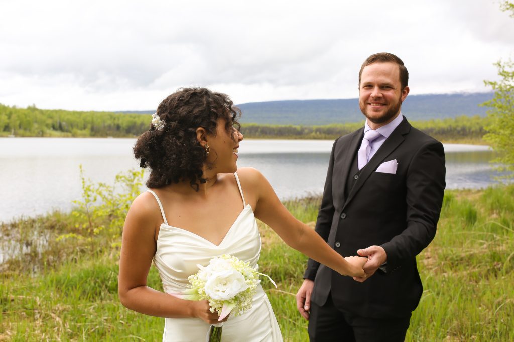 Bride holding white bouquet and groom holding hands by lakeside with mountain vista in background
