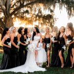 Bride and bridesmaids in black gowns with white orchid bouquets beneath moss-draped oak tree at sunset