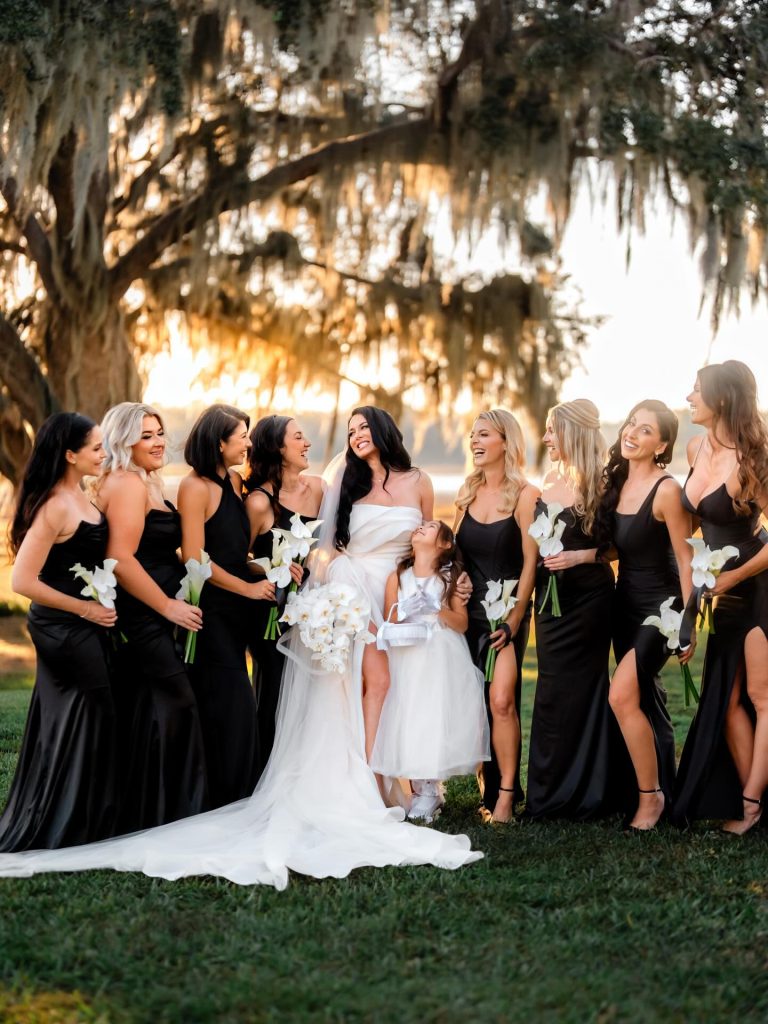 Bride and bridesmaids in black gowns with white orchid bouquets beneath moss-draped oak tree at sunset