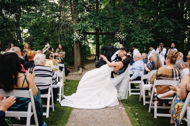 Groom dipping bride in flowing white dress during outdoor garden ceremony with guests seated along stone pathway