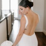Bride in elegant updo and pearl earrings poses in open-back white gown holding white bouquet