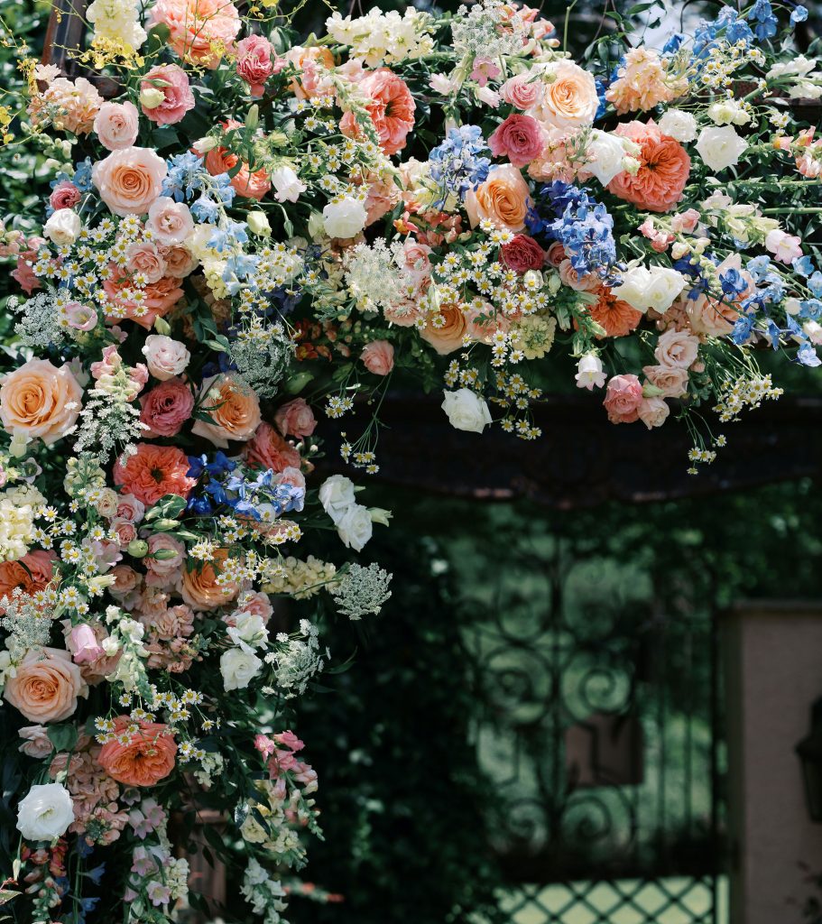 Lush floral arch featuring peach, pink, and coral roses with blue hydrangeas and white Queen Anne's lace framing ornate garden gate