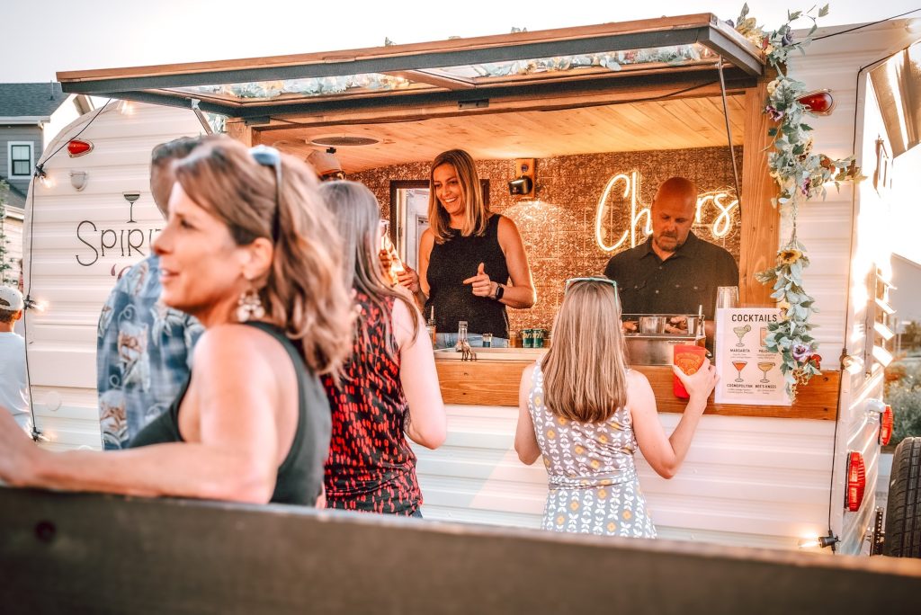 Guests gathering at vintage trailer mobile bar with "Cheers" neon sign and cocktail menu at outdoor wedding event