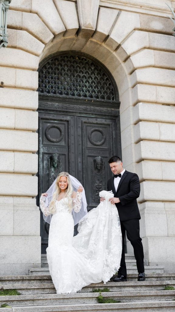 Bride and groom pose on the steps of The Ohio Statehouse in Columbus, Ohio, with the groom holding the train of the bride's lace wedding gown. The couple stands in front of the historic stone entrance during an elegant downtown wedding portrait session.