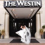 Groom carrying bride in flowing white gown and veil outside The Westin hotel entrance