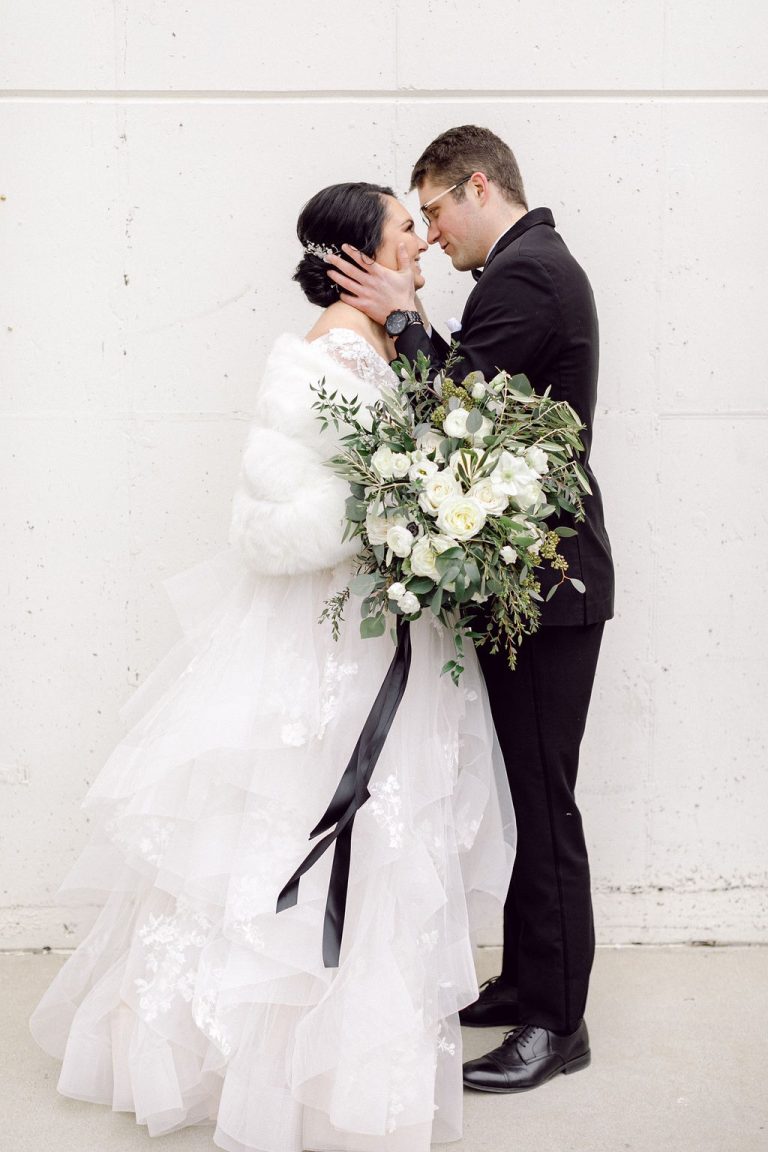 Bride and groom sharing an intimate moment against white brick wall, bride holding cascading white and greenery bouquet