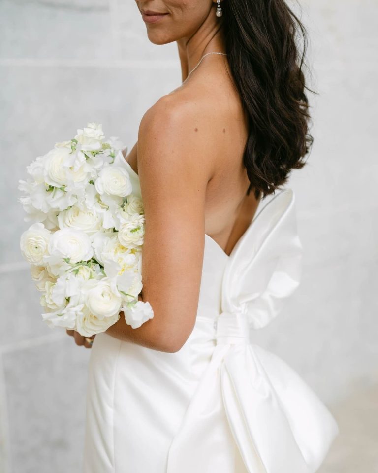 Bride in strapless white gown with bow detail holding classic white rose and ranunculus bouquet