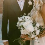 Bride holding white rose bouquet with baby's breath standing beside groom in black tuxedo with white boutonniere