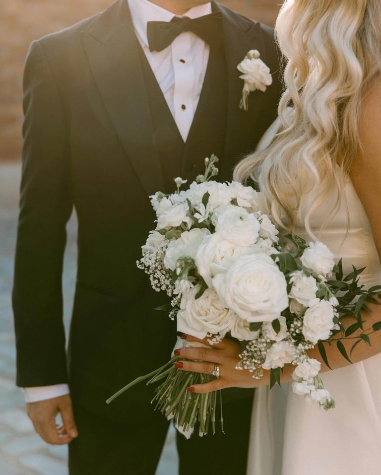 Bride holding white rose bouquet with baby's breath standing beside groom in black tuxedo with white boutonniere
