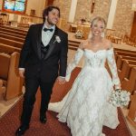 Newlyweds walking hand-in-hand down church aisle, groom in black tuxedo and bride in lace ball gown with train