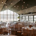 Elegant reception hall with string lights, round tables with blush linens and gold chiavari chairs, and floor-to-ceiling windows overlooking winter woods