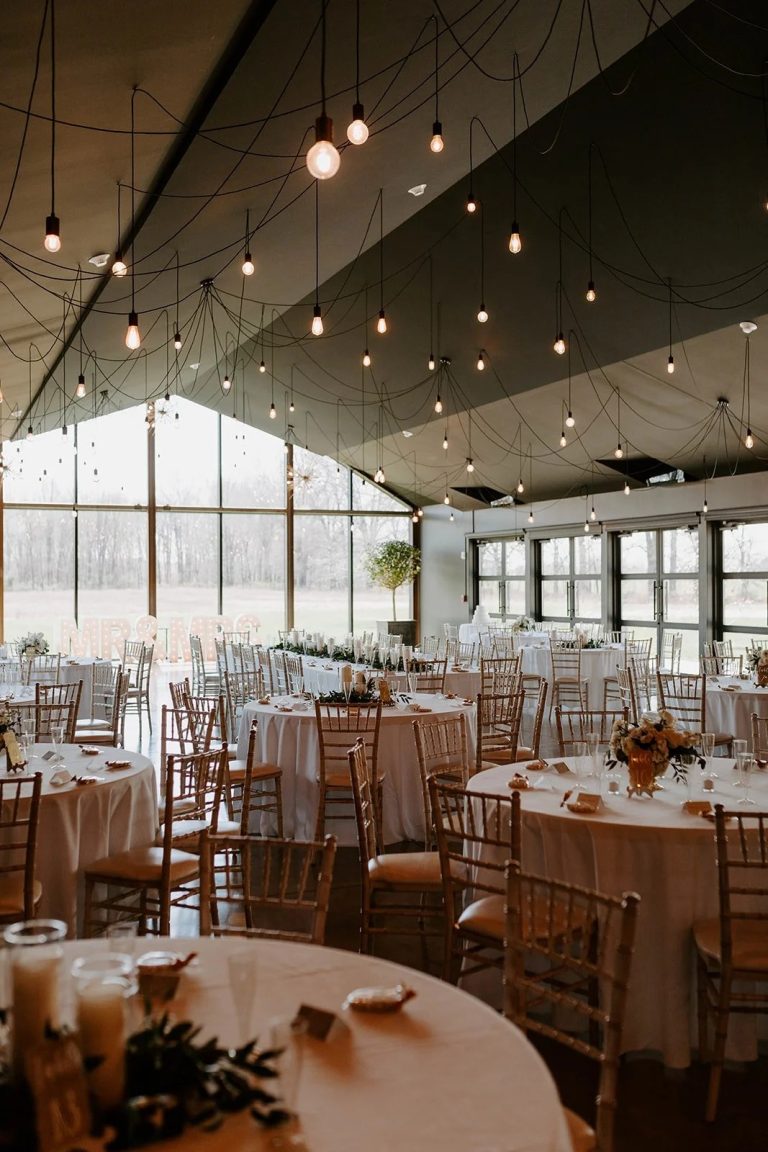 Elegant reception hall with string lights, round tables with blush linens and gold chiavari chairs, and floor-to-ceiling windows overlooking winter woods