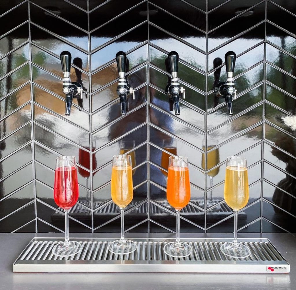 Self-serve beverage tap system with four colorful drinks dispensing against chevron tile backsplash