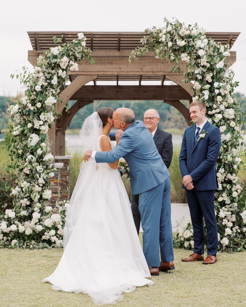 Newlyweds sharing first kiss under wooden pergola decorated with white flowers and greenery