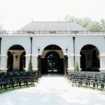White stucco building with arched colonnade and black folding chairs arranged for outdoor wedding ceremony