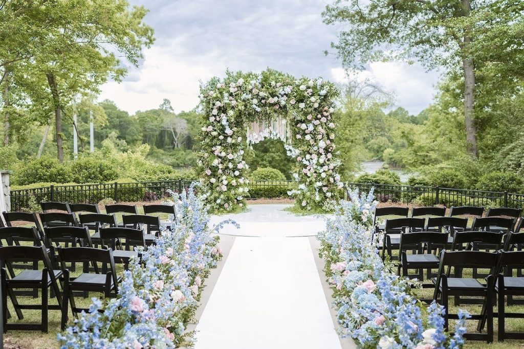 Outdoor wedding ceremony setup with floral arch and blue hydrangea-lined aisle overlooking natural landscape