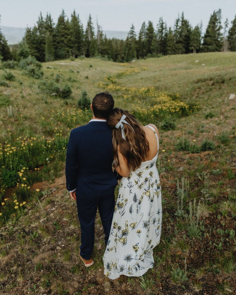 Bride and groom walking together through Colorado mountain meadow with wildflowers and pine trees