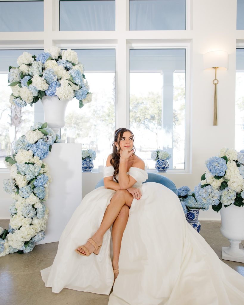 Bride in ballgown sits before blue and white hydrangea arch with matching floral arrangements in white urns