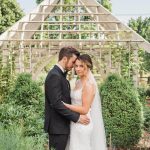 Bride and groom embracing in garden beside wooden lattice gazebo structure with lush greenery