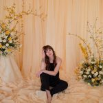Woman in black outfit sitting among cream fabric draping with yellow and white floral arrangements