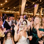 Bride and wedding guests dancing under string lights in rustic barn venue