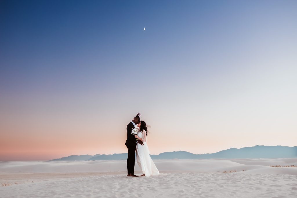 Bride and groom embrace at sunset on white sand dunes with mountains and crescent moon in background
