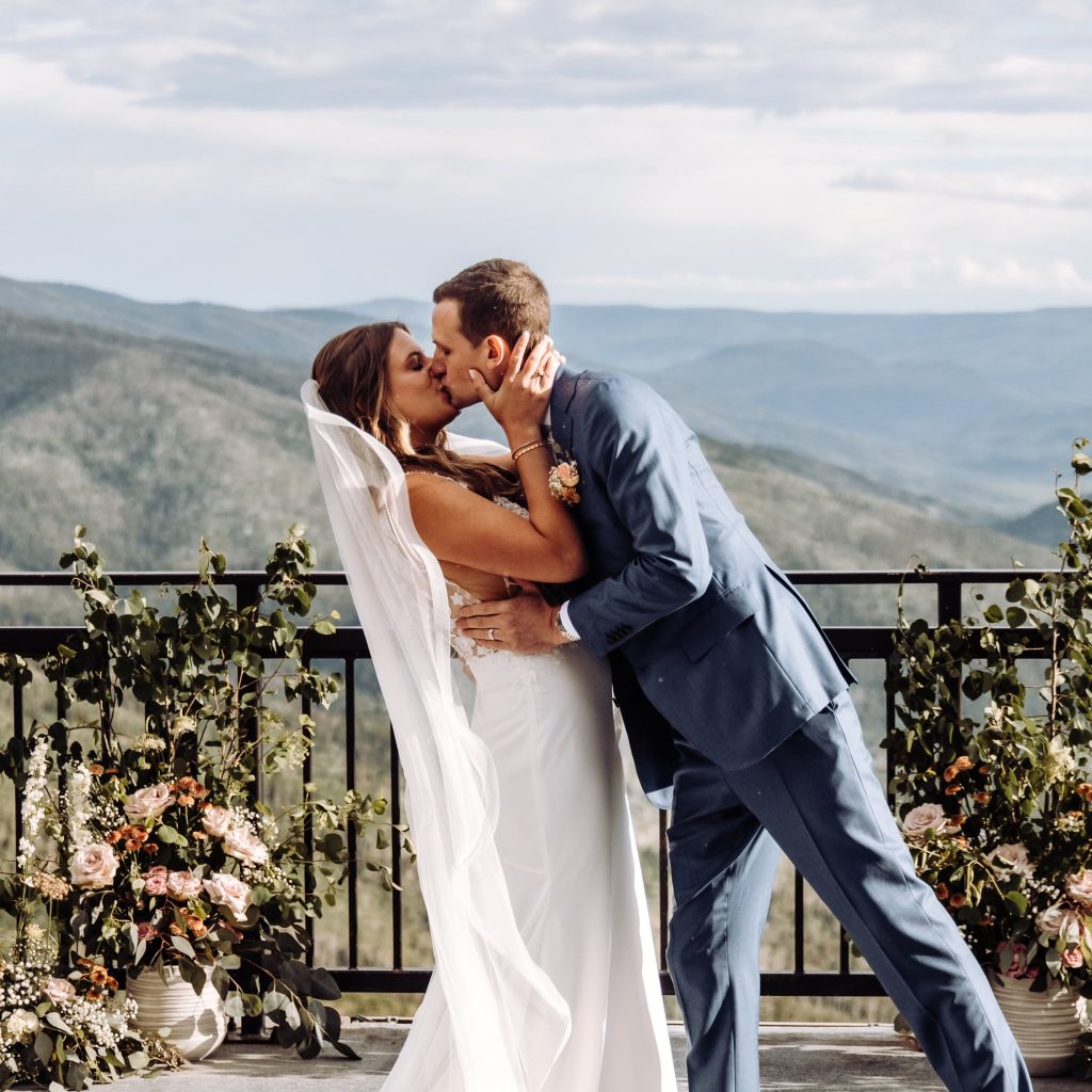 Newlyweds share first kiss at mountain overlook ceremony with floral-adorned railing and Colorado views