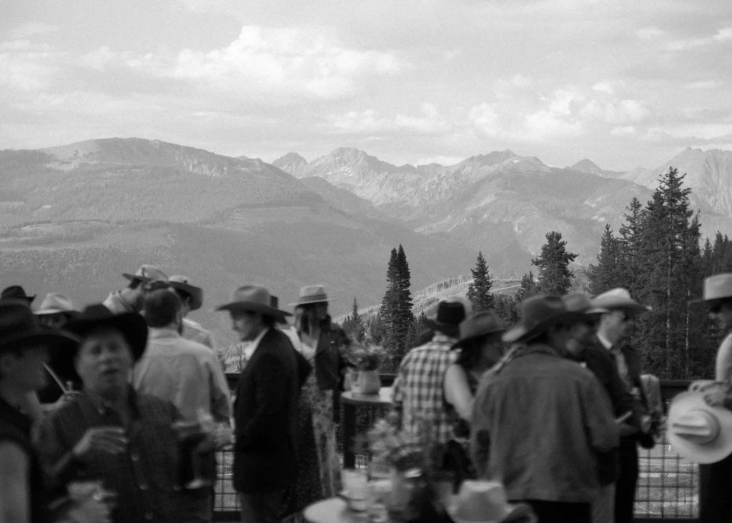 Wedding guests wearing custom cowboy hats mingle at mountain venue with scenic Rocky Mountain backdrop