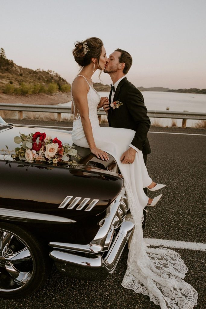 Newlyweds kiss on classic car hood at sunset overlooking reservoir, featuring bride's romantic updo with jeweled hairpiece