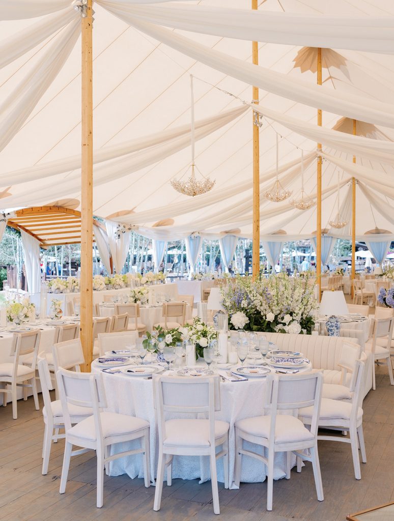 Elegant white tent wedding reception with draped fabric ceiling, crystal chandeliers, white chairs, and blue and white floral centerpieces