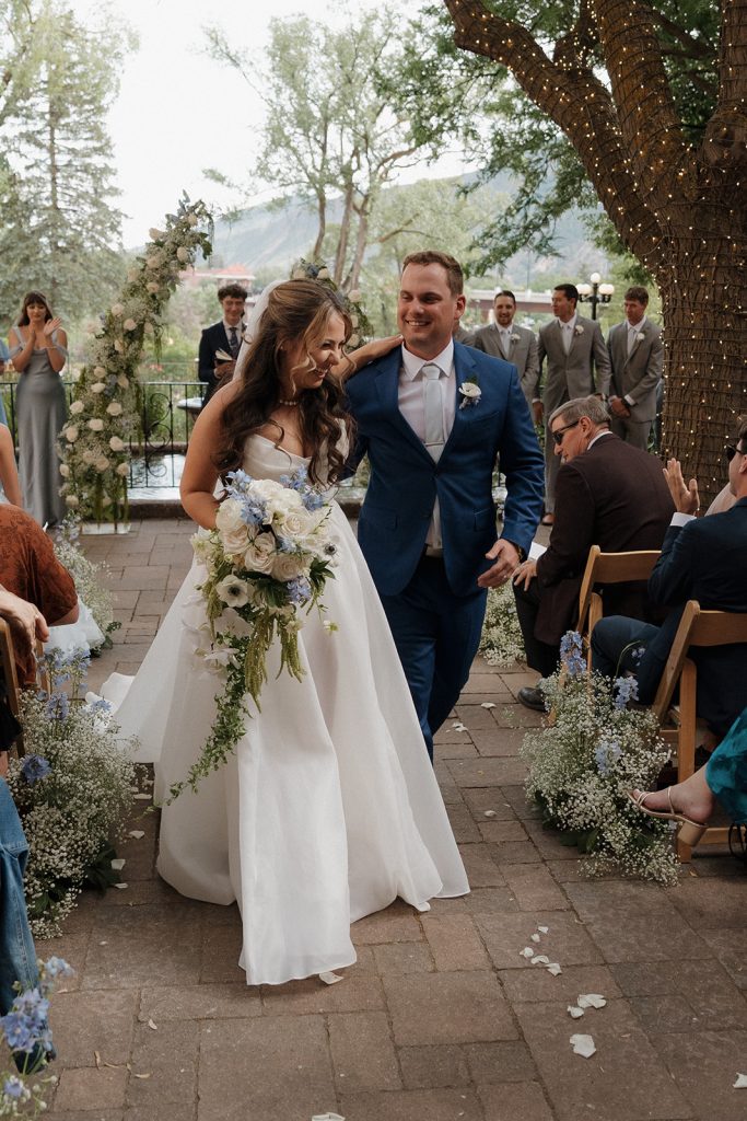 Newlyweds walking down the aisle after ceremony at outdoor Colorado mountain wedding venue with floral arch and baby's breath aisle decor