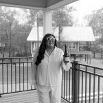 Makaela Thomas smiling on covered porch in heart-shaped sunglasses and white pajama set, holding champagne bottle