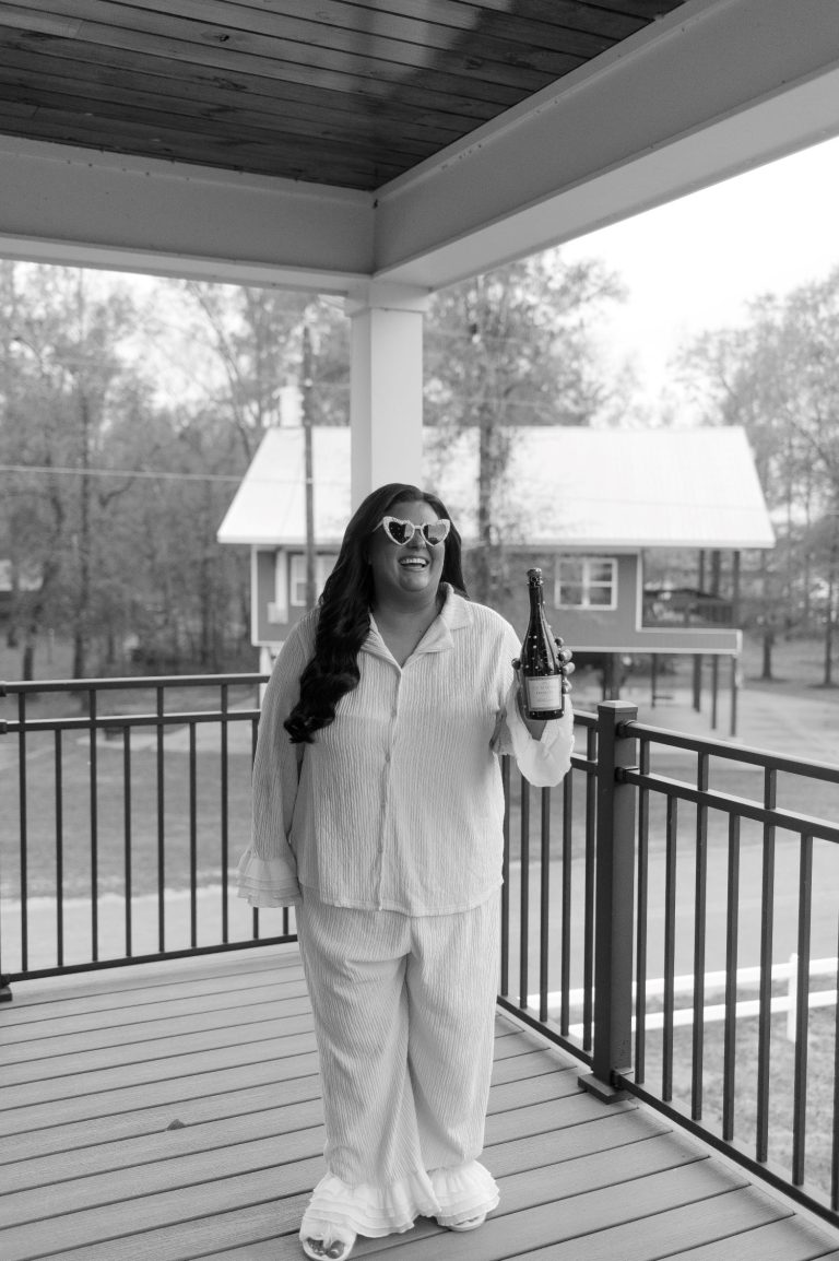 Makaela Thomas smiling on covered porch in heart-shaped sunglasses and white pajama set, holding champagne bottle