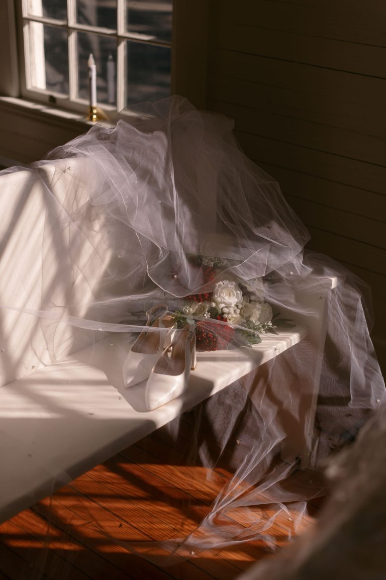 Wedding veil draped over white bridal shoes and floral bouquet in natural window light