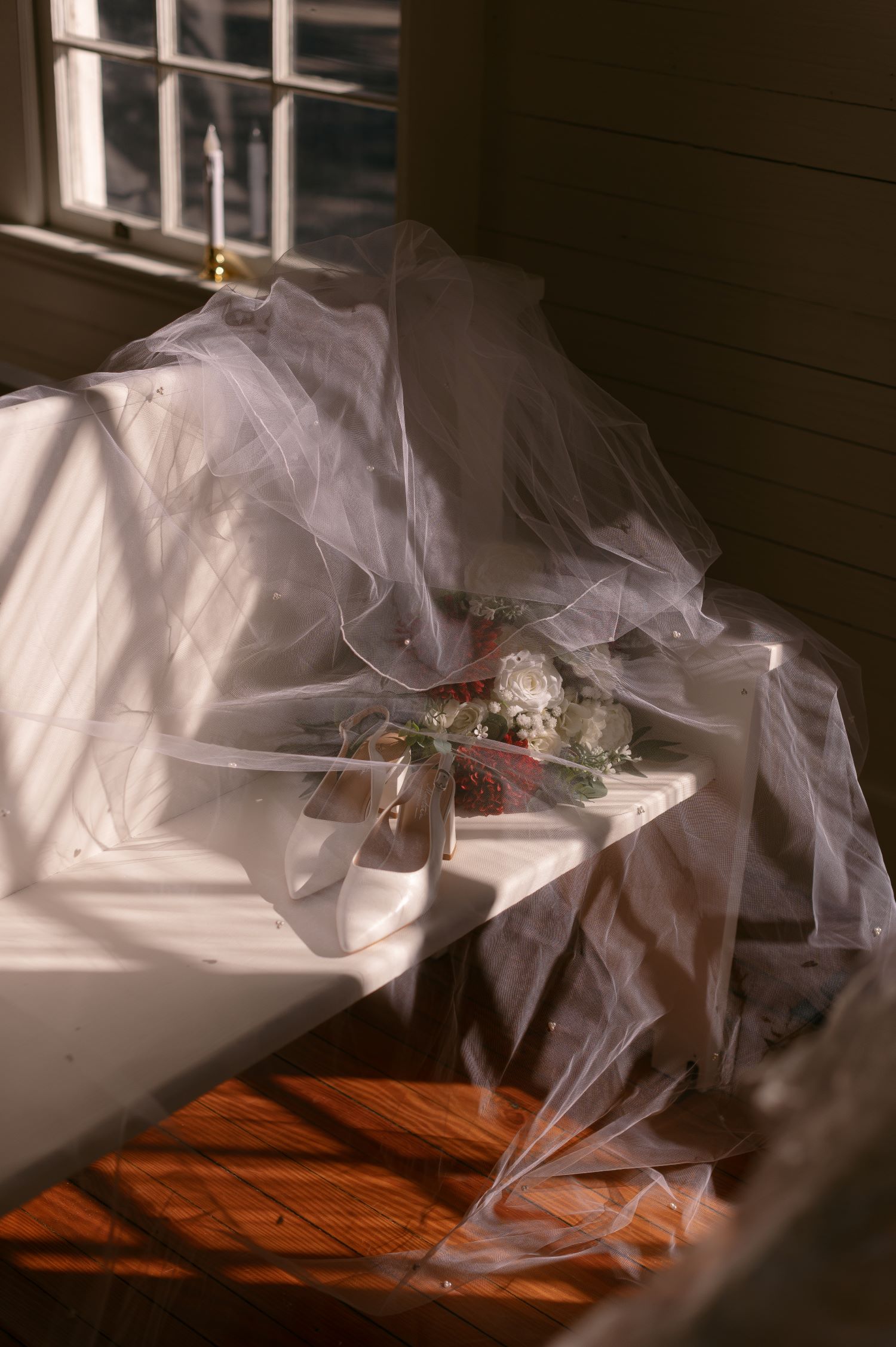 Wedding veil draped over white bridal shoes and floral bouquet in natural window light