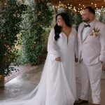 Bride and groom in matching white formal attire in rustic barn venue with string lights and greenery