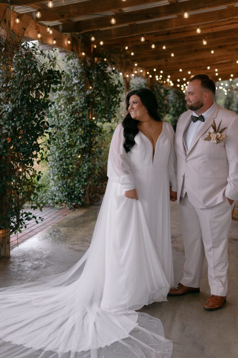 Bride and groom in matching white formal attire in rustic barn venue with string lights and greenery