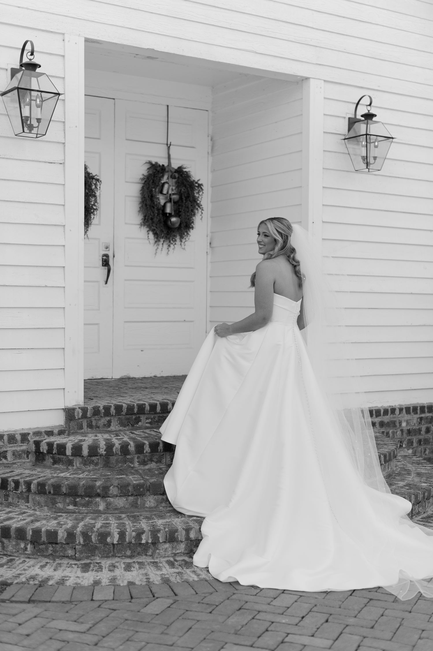 Bride in flowing white ball gown twirling on front porch with wreath-adorned door