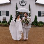 Bride and groom sharing umbrella on rainy wedding day in front of white barn venue