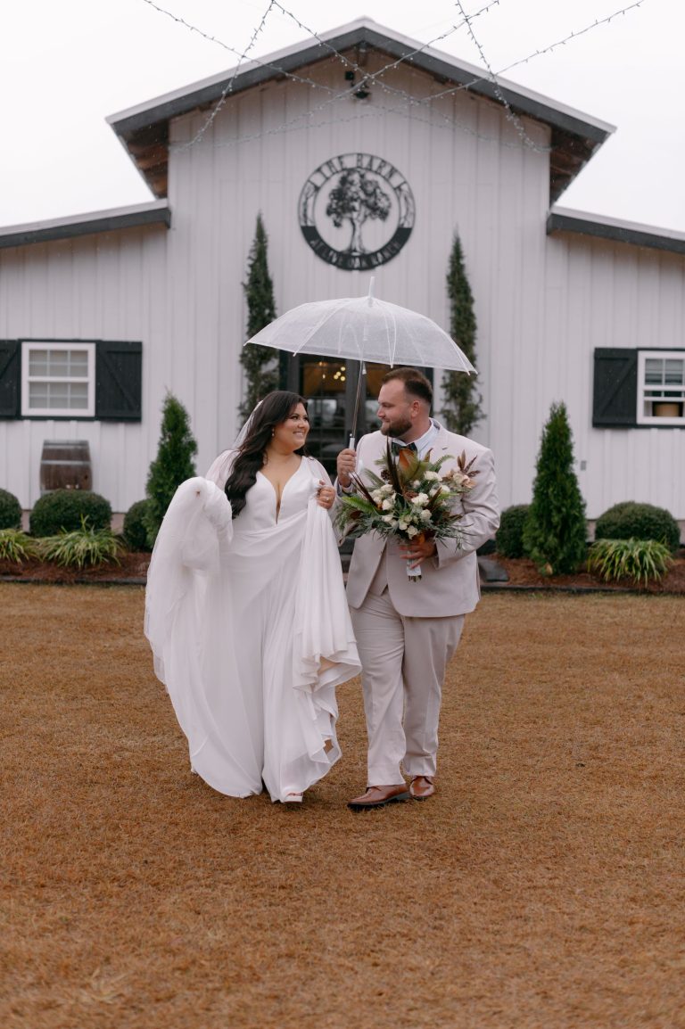 Bride and groom sharing umbrella on rainy wedding day in front of white barn venue