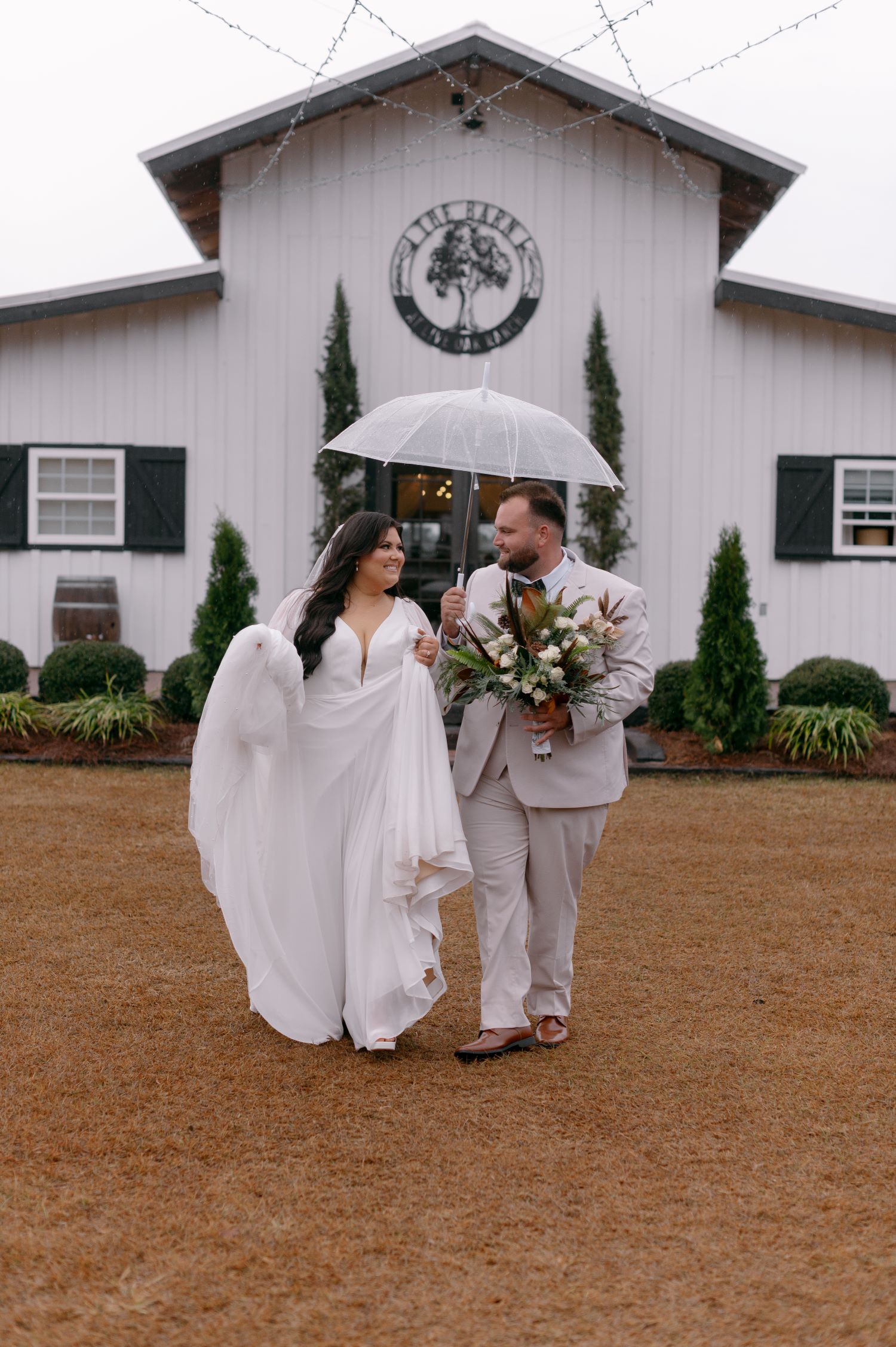 Bride and groom sharing umbrella on rainy wedding day in front of white barn venue