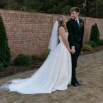 Newlyweds holding hands and gazing at each other near brick wall and manicured garden