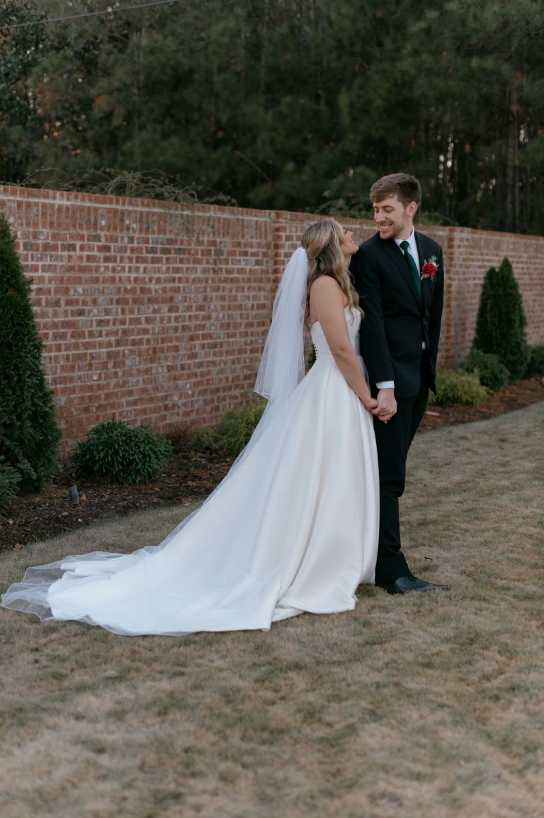 Newlyweds holding hands and gazing at each other near brick wall and manicured garden