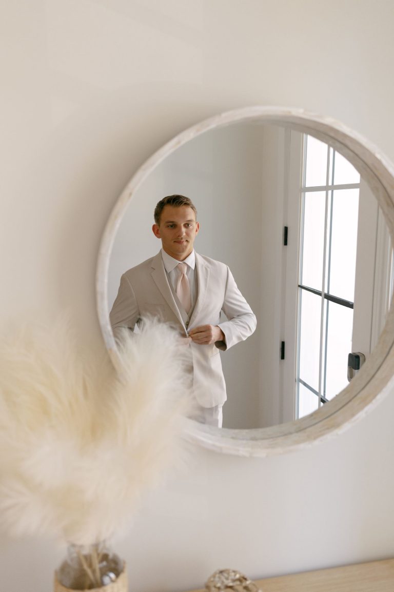 Groom in tan suit and blush tie reflected in round mirror with pampas grass in foreground