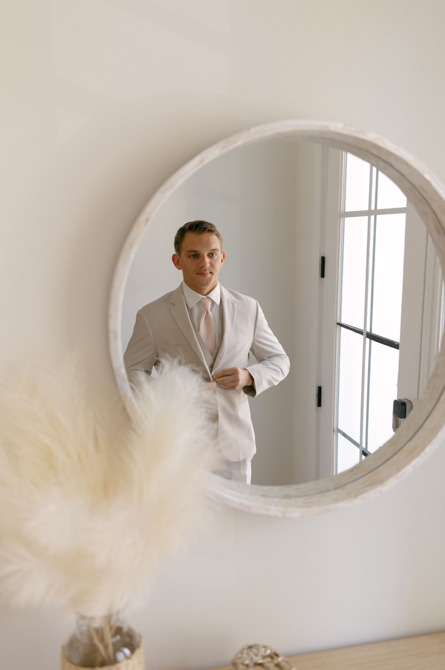Groom in tan suit and blush tie reflected in round mirror with pampas grass in foreground
