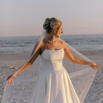 Bride in strapless ballgown with pearl-adorned veil spinning on beach during golden hour