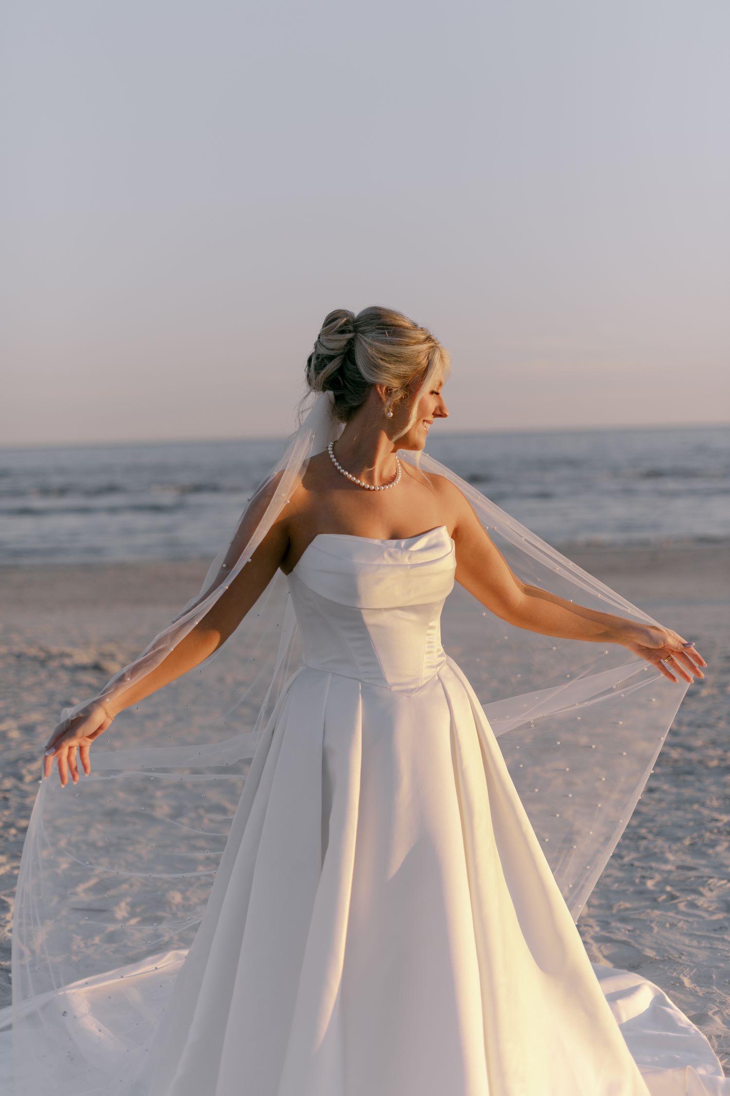Bride in strapless ballgown with pearl-adorned veil spinning on beach during golden hour