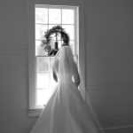 Bride in veil and gown silhouetted before window with wreath in shiplap room