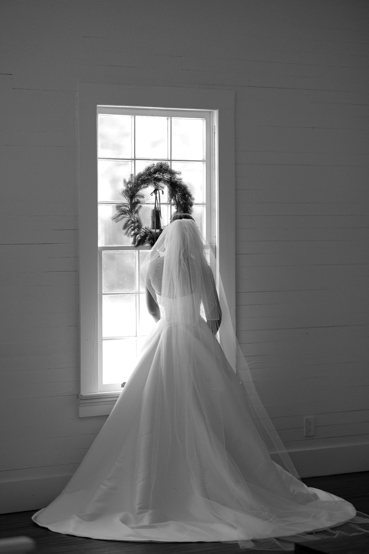Bride in veil and gown silhouetted before window with wreath in shiplap room